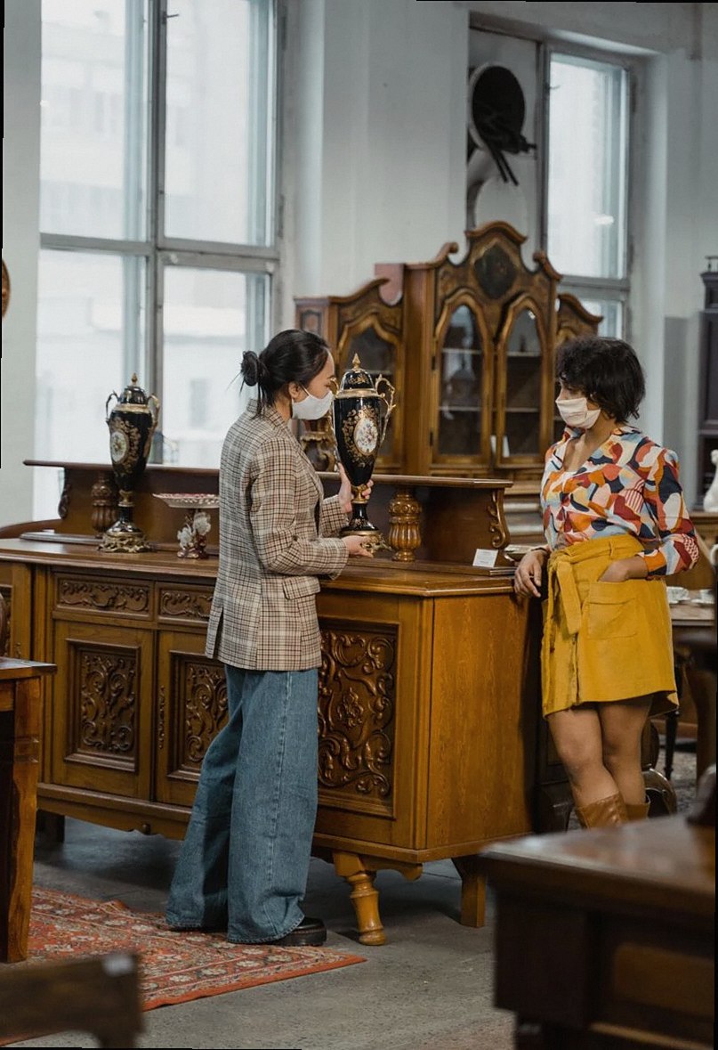 Two women wearing face masks browsing furniture in a vintage shop indoors.